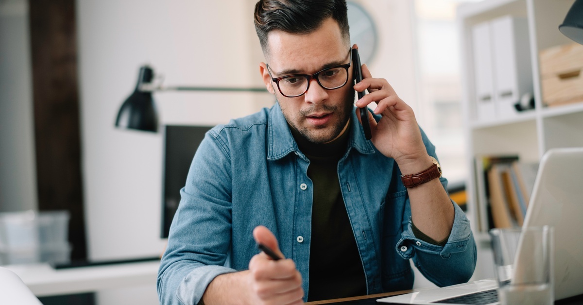 A man in a button-down shirt and glasses sitting at a desk and holding a cellphone to his ear as he looks downward.