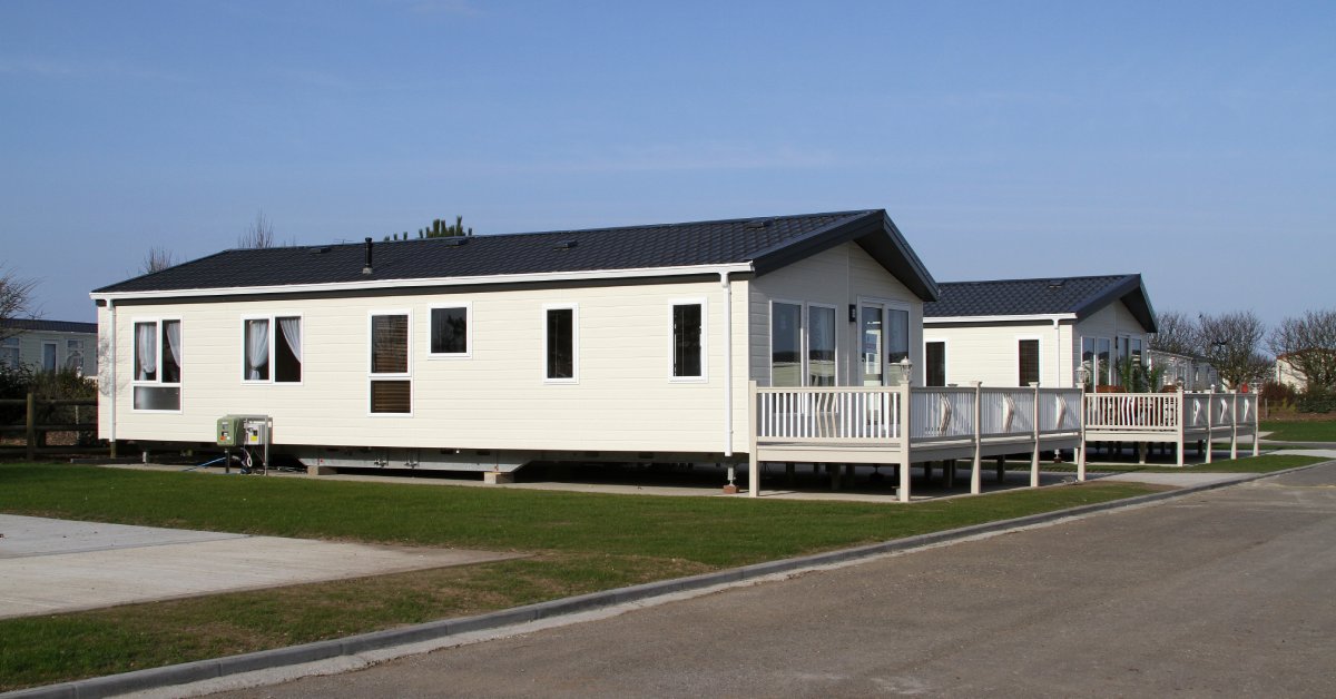 A small white home with a black roof and front porch sits on short stilts the edge of a property near the street.