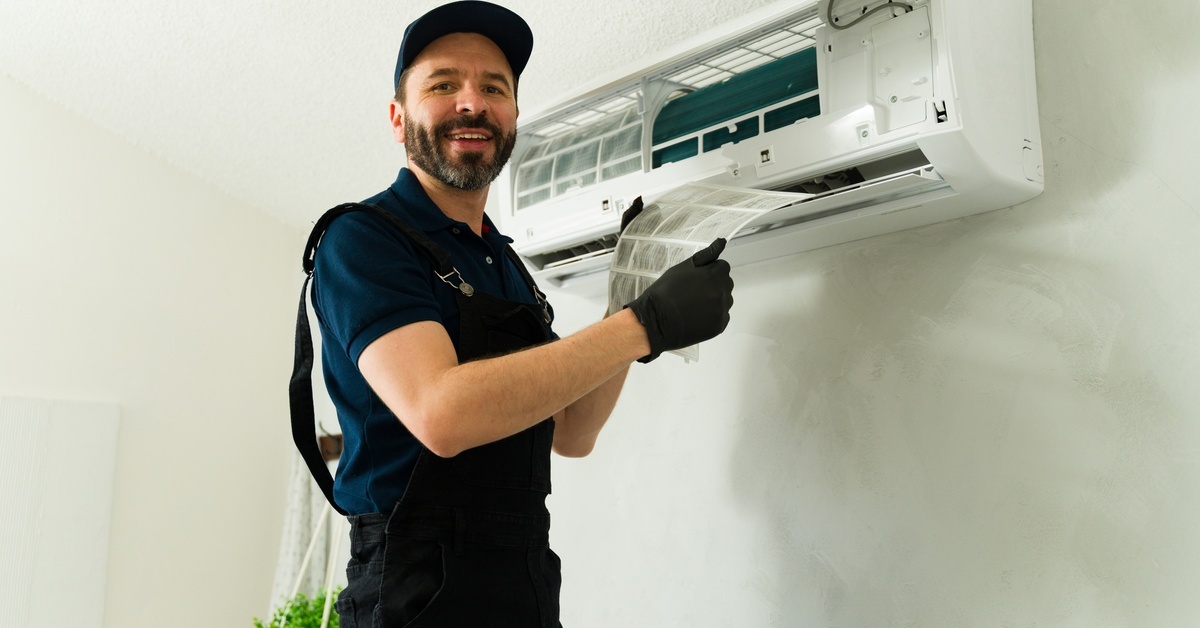 A man wearing a black shirt, pants, and hat holds the filter from a wall-mounted split system AC unit inside a home.
