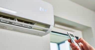 A hand holding a small remote and pointing it upward towards a wall-mounted split-system AC unit that has the number 21 on it.