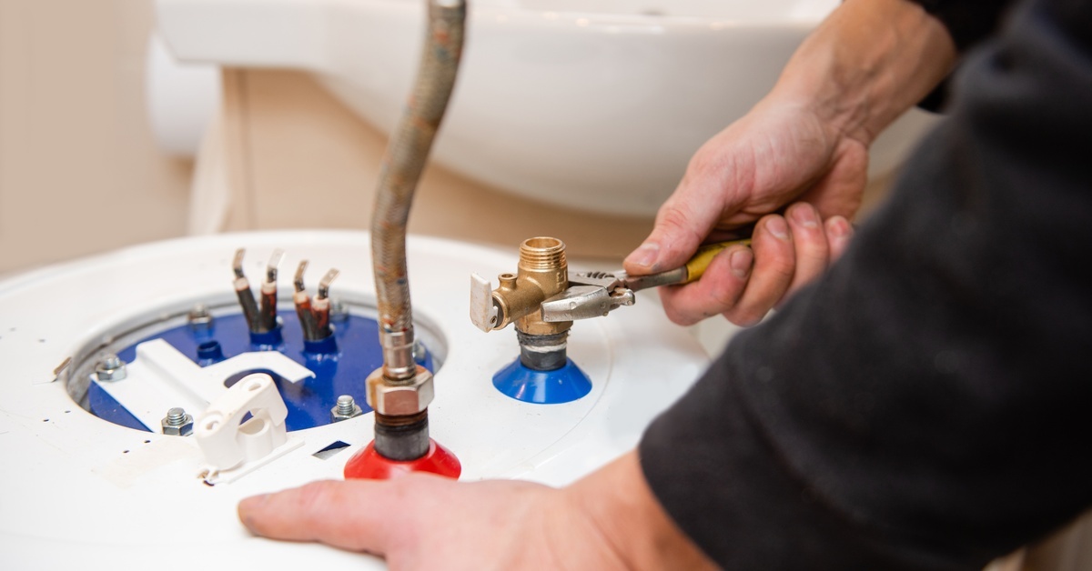 Someone in a black long sleeve shirt holding a wrench to the cold water valve on a water heater. There’s a pipe connected to the hot water valve.