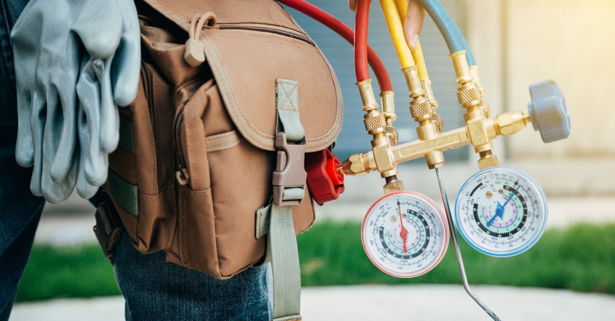 A hand holding a heating and cooling tool with hoses connected to the device; a house is slightly blurred in the background.