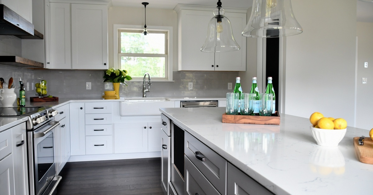 A modern kitchen contains white cabinets, a white apron sink, a large island, white countertops, and pendant lighting.