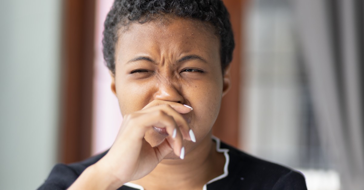 A close-up view shows a woman with short hair holding her hand under her nose. She has a disgusted look on her face.