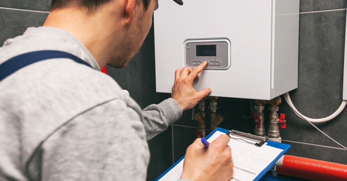 A man wearing blue overalls and a black baseball cap is pressing the button on a white water heater and holding a clipboard.