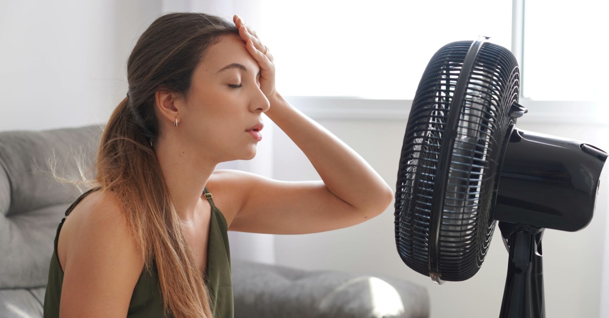 A young woman wearing a green shirt sits in front of a black fan. Her eyes are closed, and her hand is on her forehead.