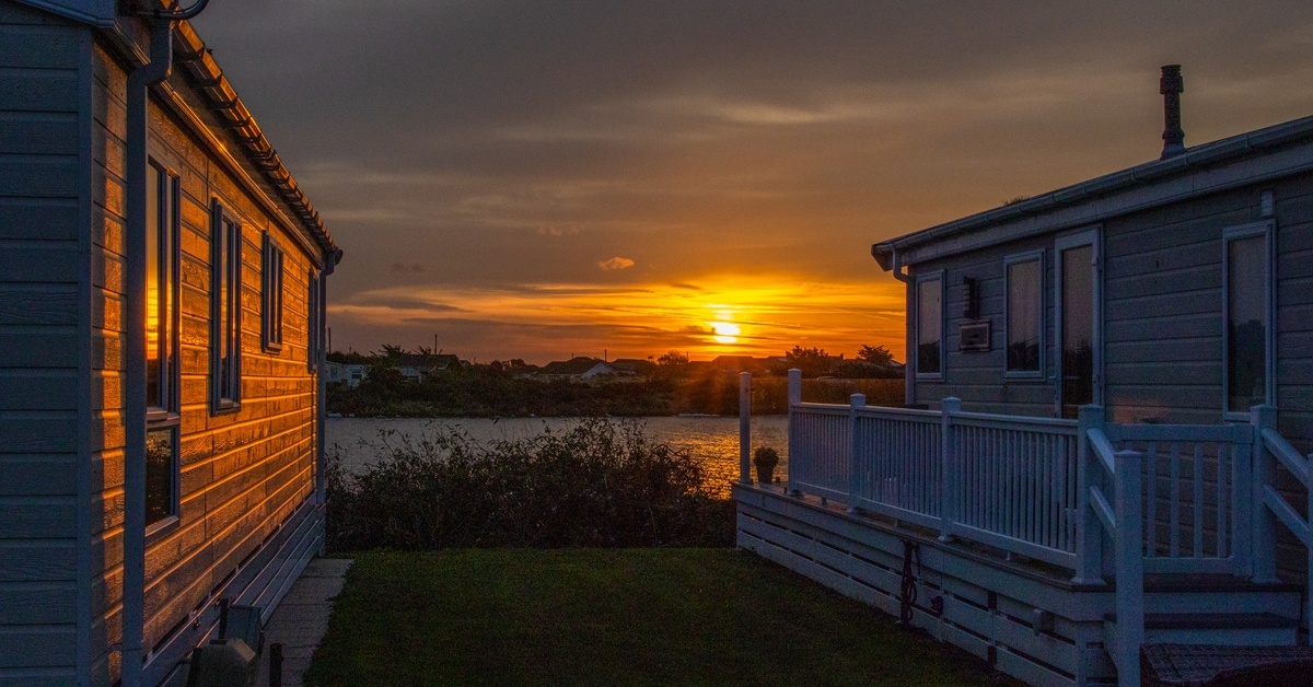 The sun is setting between two mobile homes that sit next to a large body of water. Clouds are in the sky.