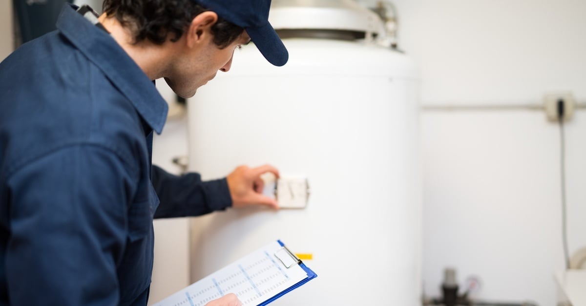 A man wearing a blue shirt and a baseball hat is holding a clipboard and examining a white hot water heater.