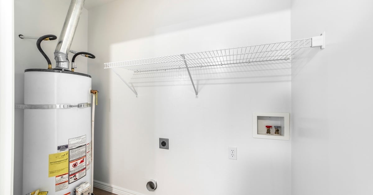 A white hot water heater sits inside a utility room with white walls, laundry hookups, and a white wire shelf.
