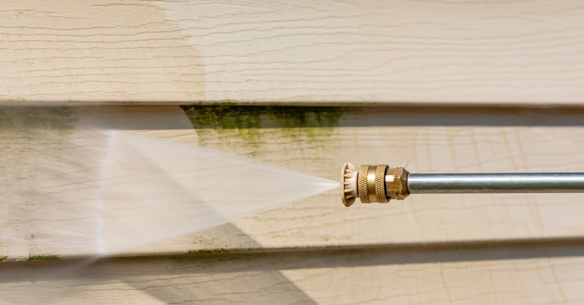A close-up view shows a metal pressure washer with a copper tip spraying water onto to tan siding to remove the mildew.
