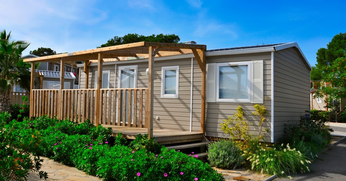 A small mobile home has tan siding and a cute wooden patio attached to the side. There's landscaping around the home.