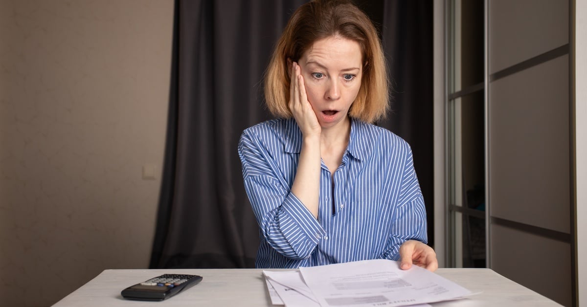 A woman sits at a white table with a shocked expression on her face. She's looking at paperwork with one hand on her cheek.