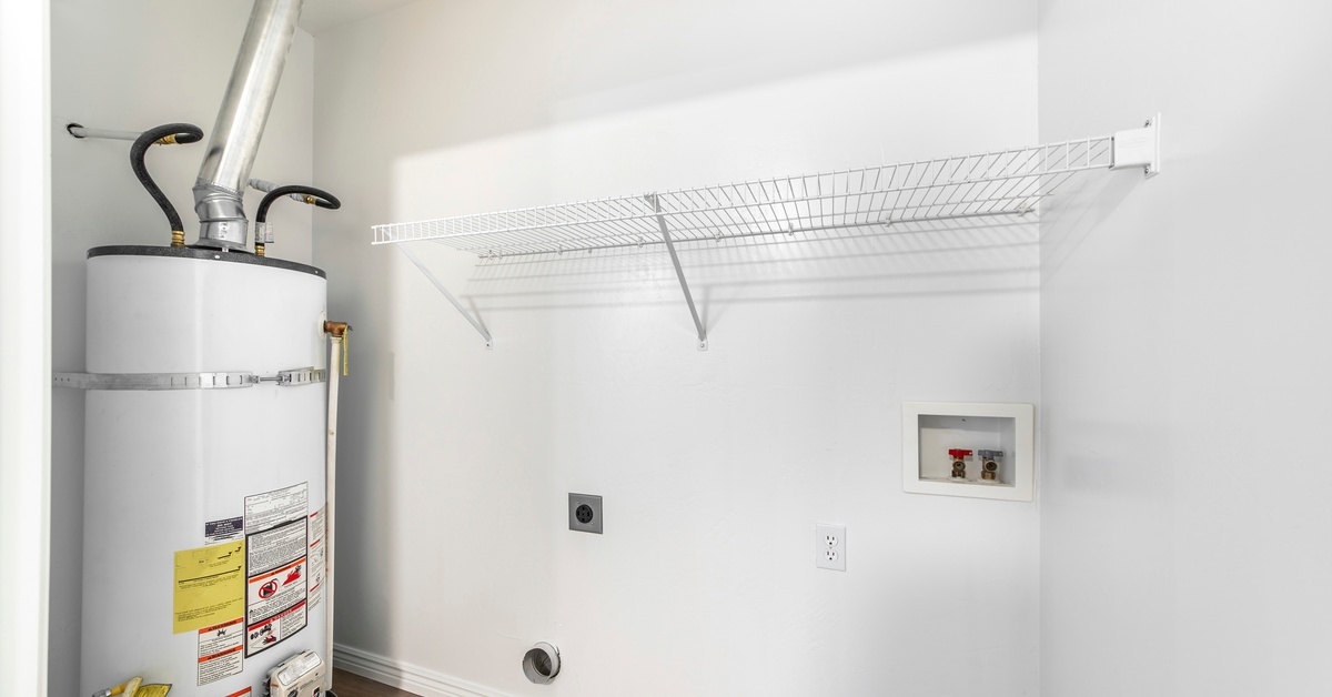 A white water heater sits alone in a white closet that contains the hookup for a washer and dryer and a white rack.