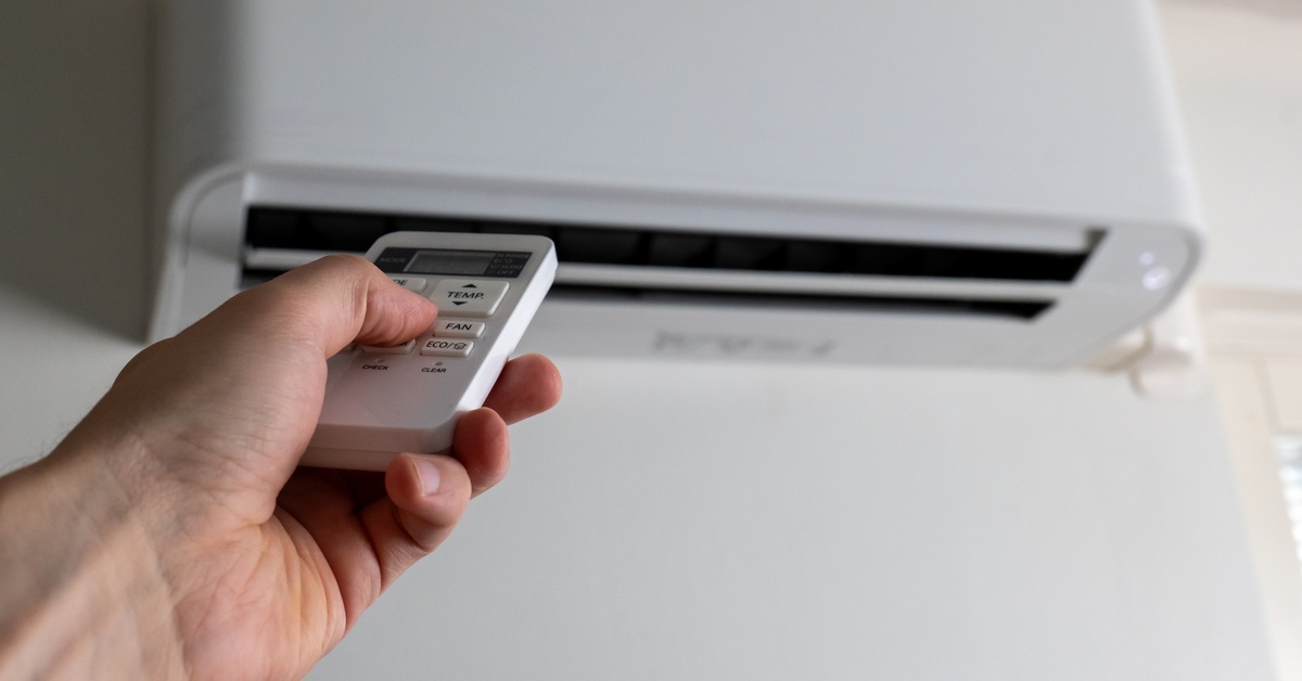 A close-up view shows a person holding a silver remote and pointing it at a ductless AC unit that sits on a wall.