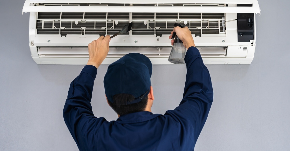 A technician wearing a jumpsuit and a blue hat is using a spray bottle and a black, plastic scraper to clean a mini split AC.