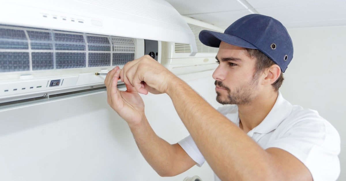 A man wearing a white polo shirt and a blue baseball hat is using a screwdriver to fix a mini split AC system.