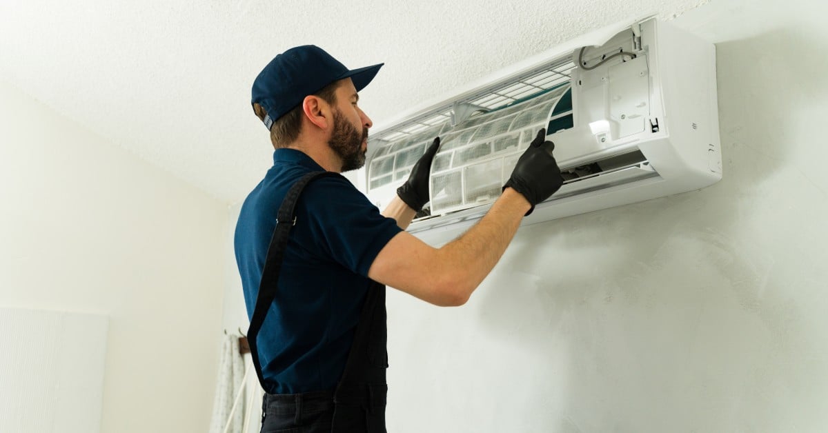 A man wearing a blue shirt, black pants, and a baseball hat is working on a white mini split system.