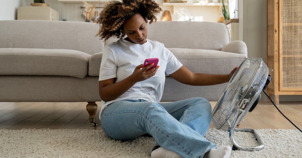 A woman sits on the floor on a rug in front of a gray couch. She's scrolling on her phone with an arm resting on a fan.