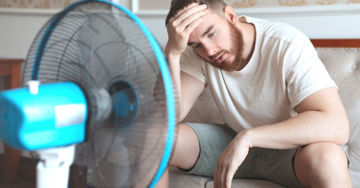 A man sits on a leather couch with his hand on his forehead. An electric fan is in front of him, cooling him off.