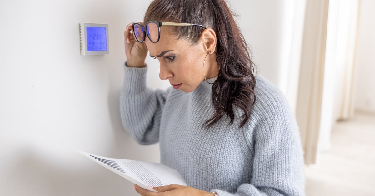 A woman wearing a gray sweater holds a piece of paper and lifts up her glasses to look at it. She looks concerned.