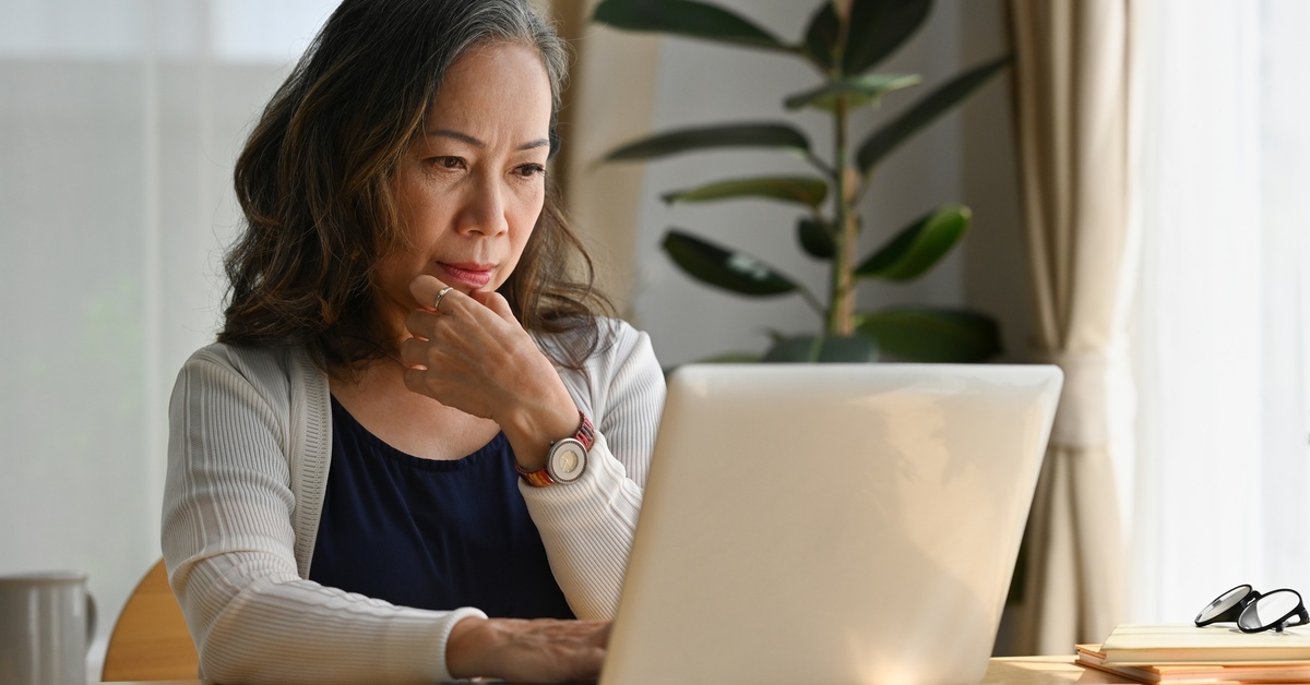 A woman sits at a wooden table looking at an open laptop. She has one hand resting on her chin and the other on the keyboard.