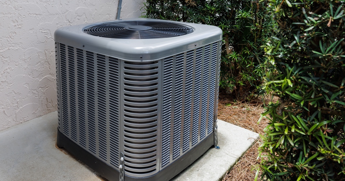 A large, dark-gray air conditioning unit sits on a concrete slab surrounded by lush, green bushes.