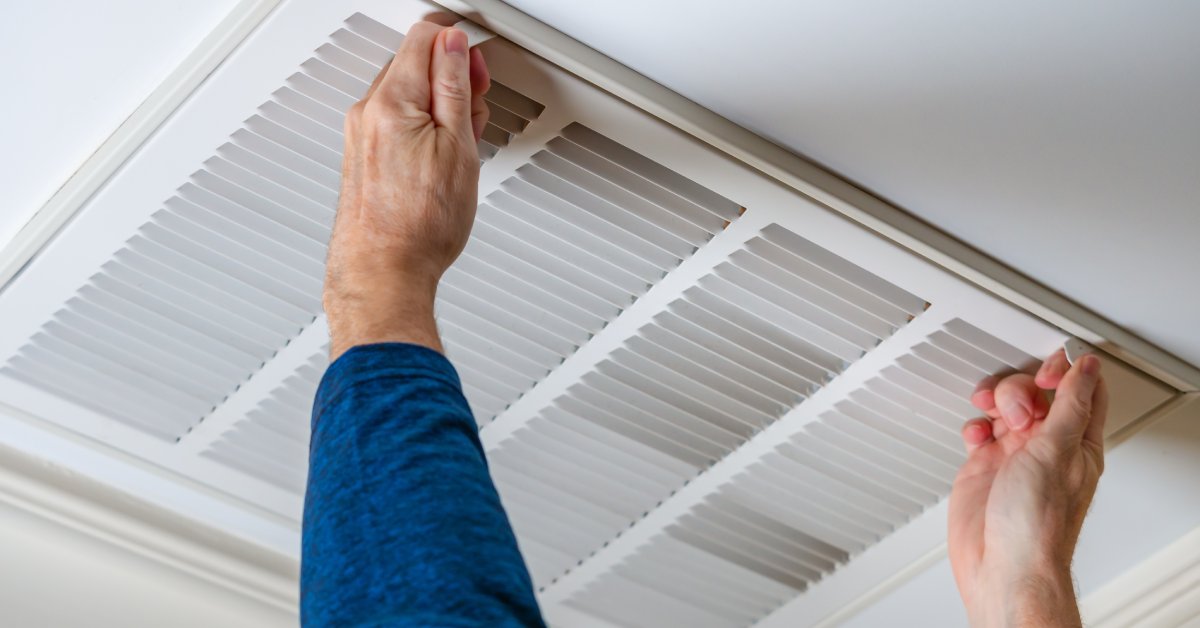  A close-up view shows a person wearing a blue long-sleeve shirt pressing the tabs on a white ceiling air vent.