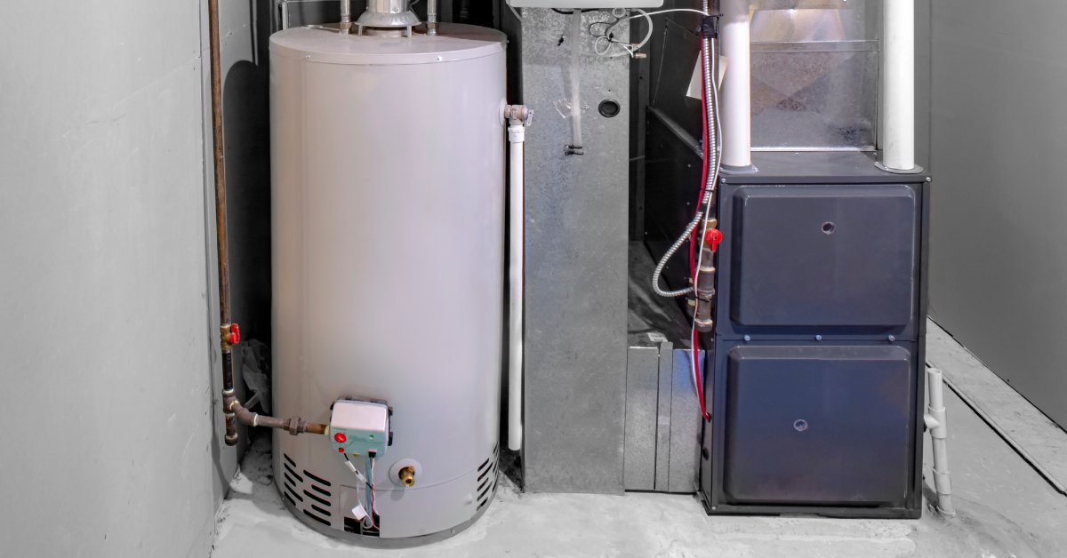 A large off-white tank water heater system with pipes connected to it sits on the floor beside an air duct and HVAC system.
