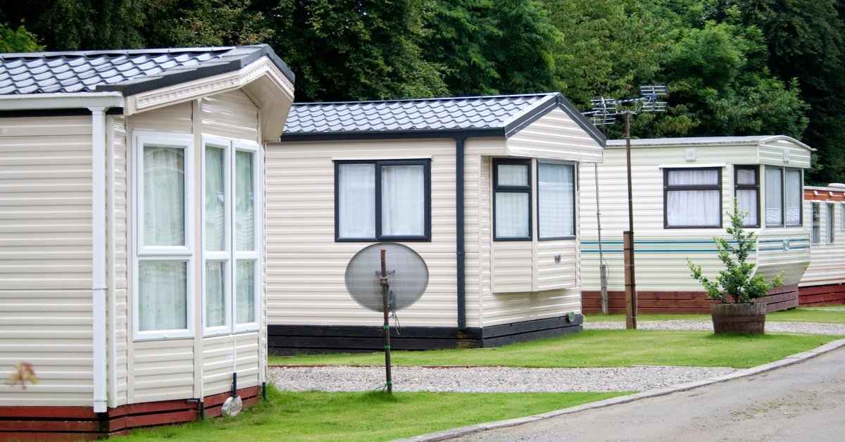 A row of white mobile homes in between an unpaved road and a line of trees. Some have satellite dishes in the front.