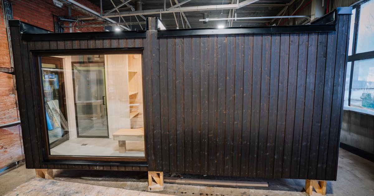 A section of a house sitting on blocks inside of a building. The structure has a glass door, windows, and dark wood exterior.