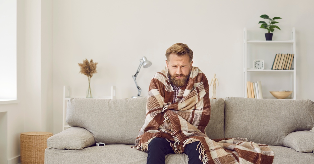 A man sitting inside a home on a gray couch with his eyes shut. He has a brown plaid blanket wrapped around him.