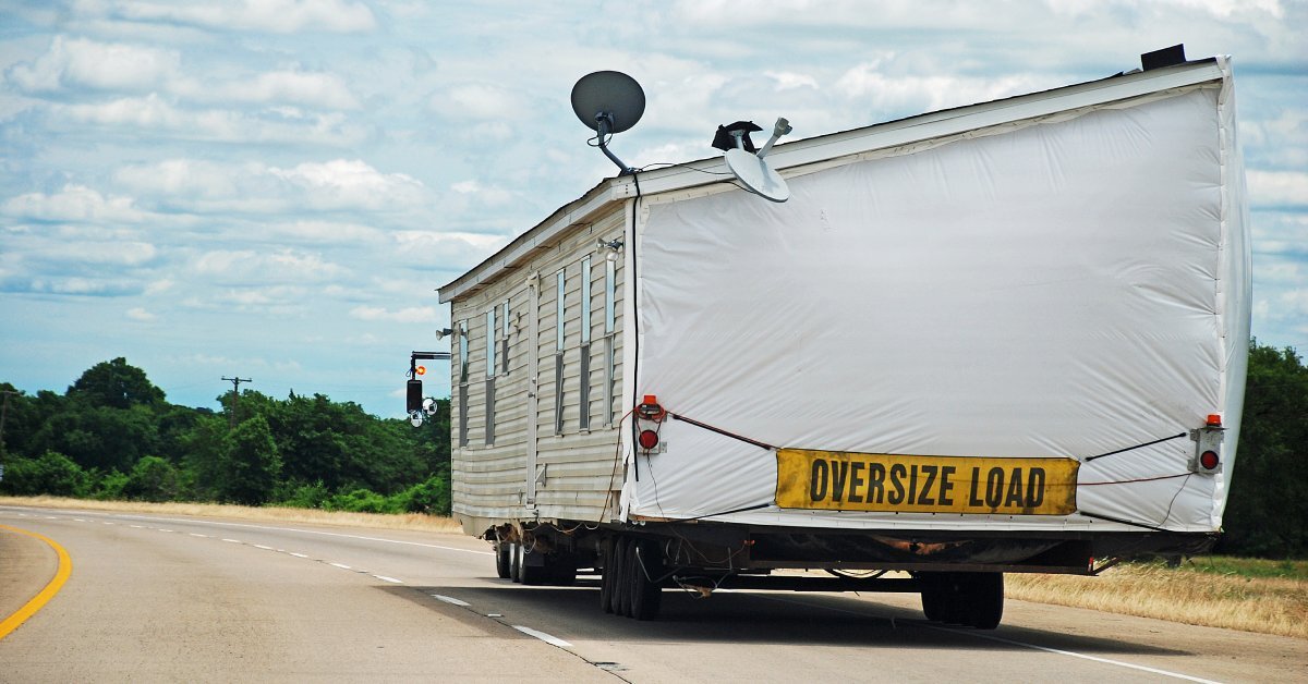 A white house on the flatbed of a truck driving down an open road. There’s a sign labeled “oversize load” on the house.