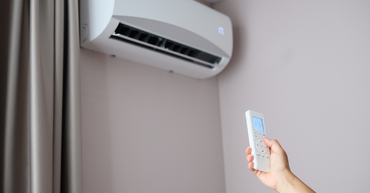 A hand holds a white remote and points it upward toward a split system AC unit mounted on a wall beside a gray curtain.