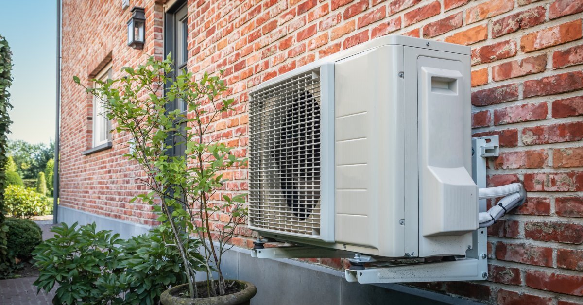 A heat pump AC unit mounted to the brick exterior wall outside of a building. There’s a potted plant by the heat pump.