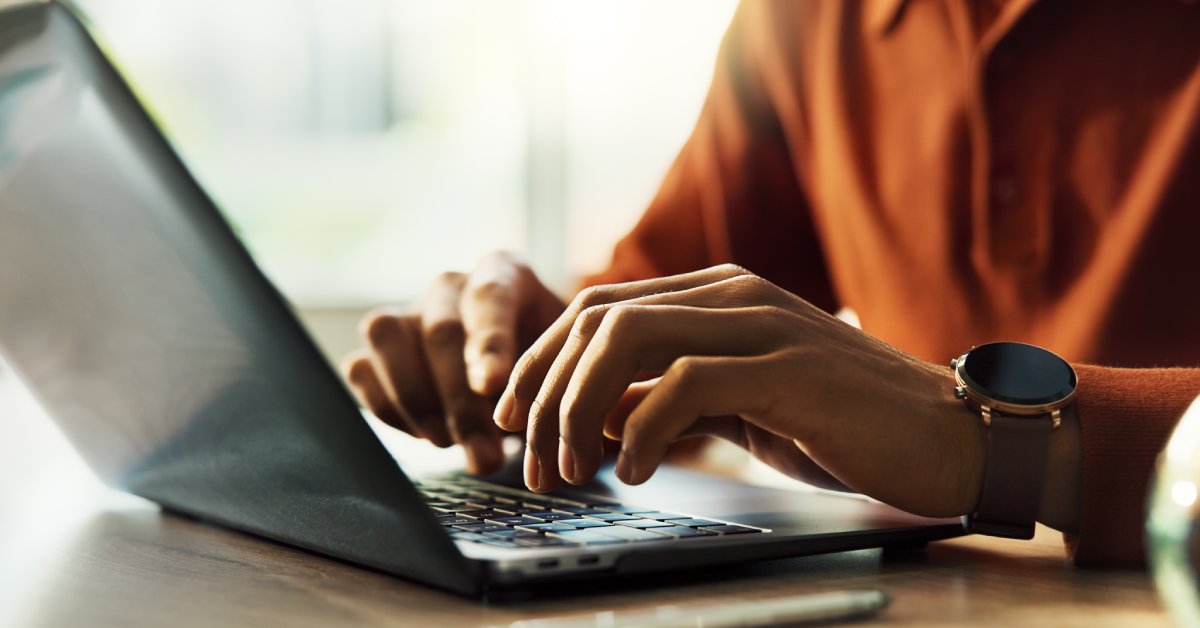 A person’s hands hover above the keyboard of a laptop that’s sitting on a wood surface. The person has an orange shirt on.
