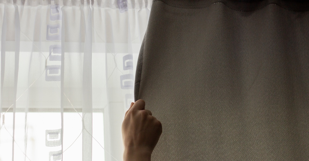 A hand pulling a tan curtain to open or close it. There’s a sheer white curtain pulled shut in front of the window.