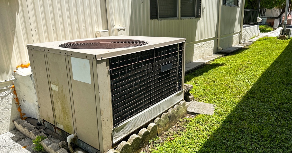 An HVAC unit sitting outside of a white, manufactured home with black shutters that has awnings over the windows.