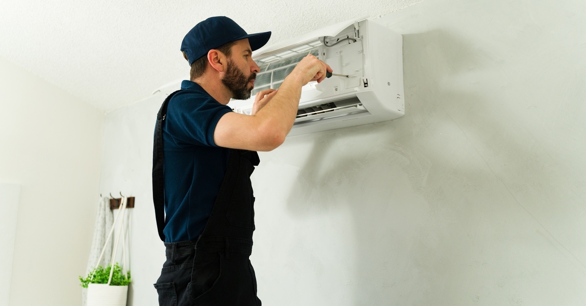 A man standing near a split-system AC unit that has the front panel open. The man is holding a screwdriver and working on it.