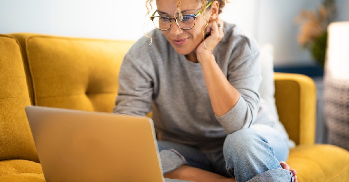 A woman wearing glasses sits on a yellow couch and leans forward to look at the laptop sitting on the cushion beside her.