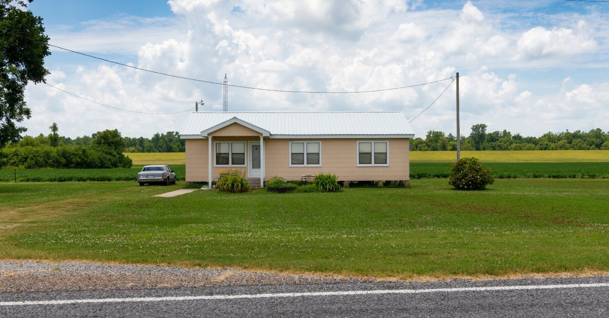 A cream-colored manufactured home sitting alone in a field with a car beside the home. Lush, green grass surrounds the home.