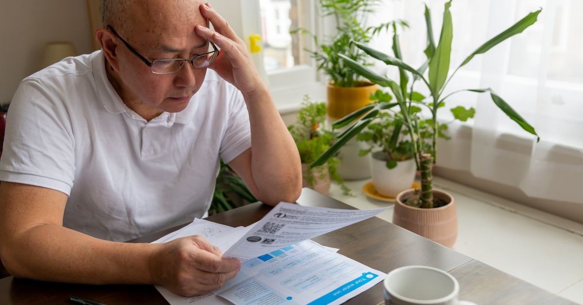 A man in a white T-shirt and glasses sitting at a table with papers on it. He's holding a paper with one hand and his other hand is on his head.