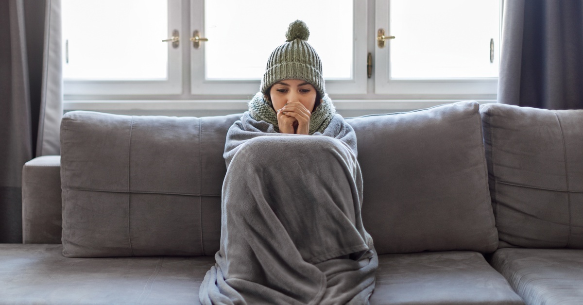 A woman inside sitting on a gray couch in front of a window. She’s wearing a hat and has a blanket over her lap.