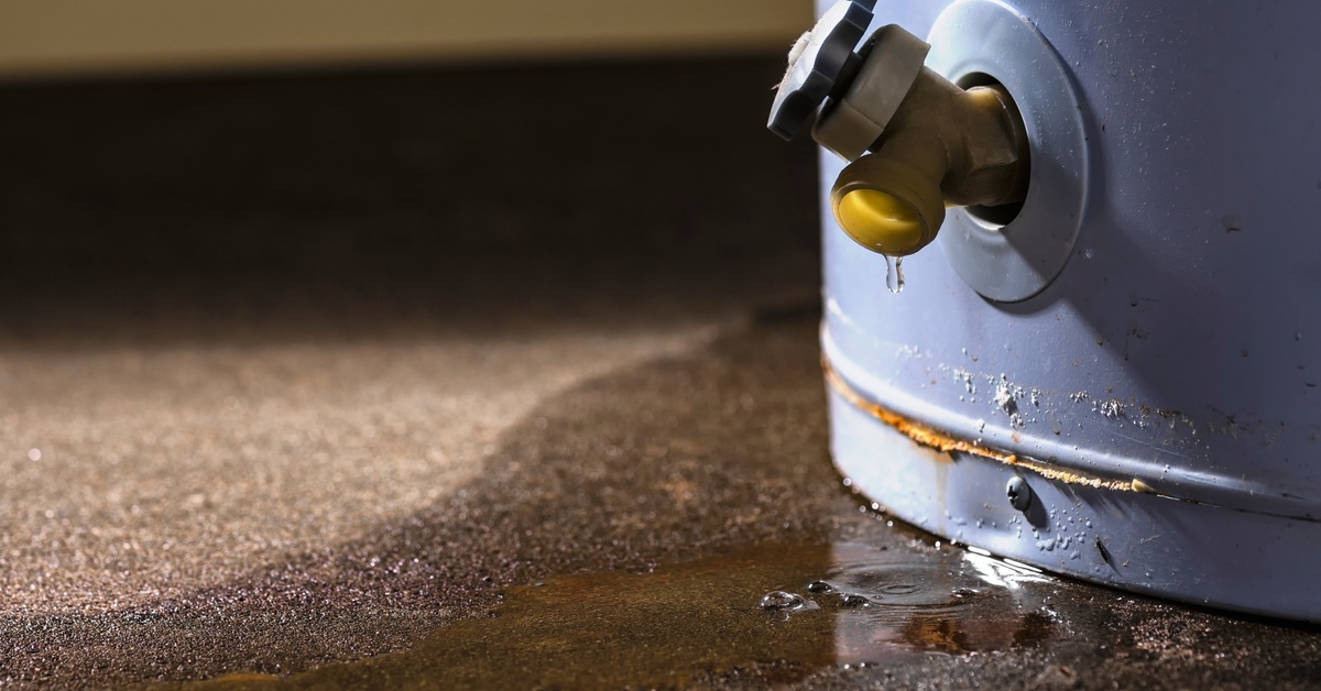 A closeup of the base of a water heater with water dripping from the spout; there’s a small puddle surrounding the unit.