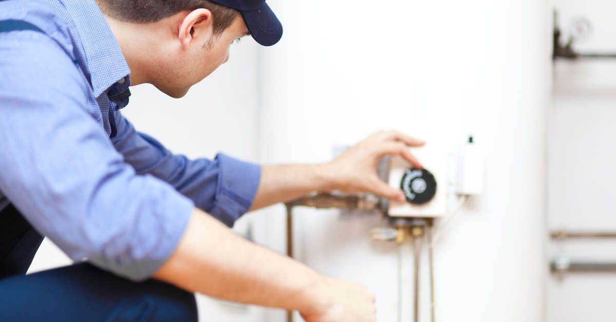 The back side of a man in a blue shirt that has the sleeves rolled up is kneeling down by a water heater with his hand on the dial.