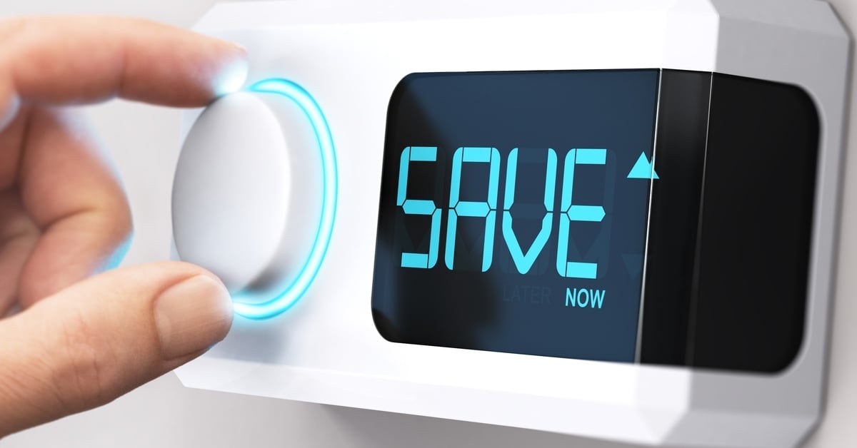A hand on the dial of a wall-mounted thermostat; the screen of the thermostat says “SAVE NOW’ in blue font.