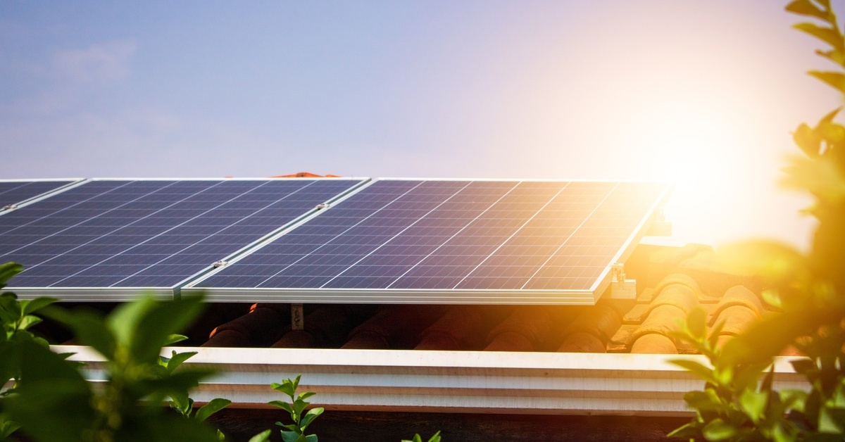Solar panels mounted to the roof of a home; the sun shines brightly in the background and greenery is in the foreground.