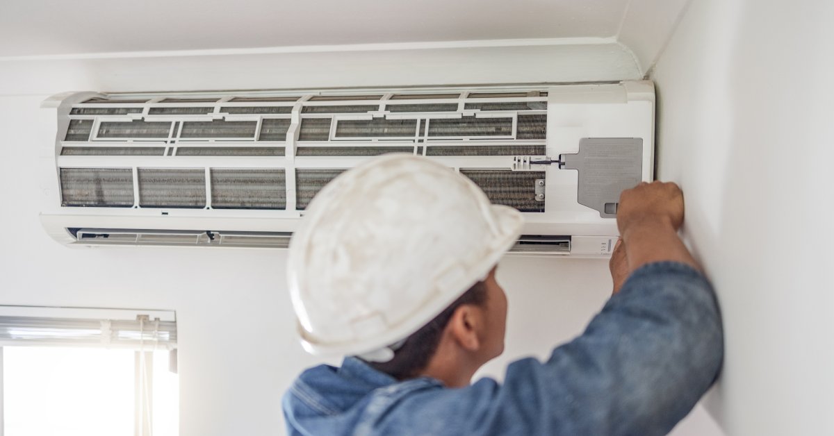 The backside of a man in a long-sleeved blue shirt and white hard hat standing by an indoor, wall-mounted mini-split.