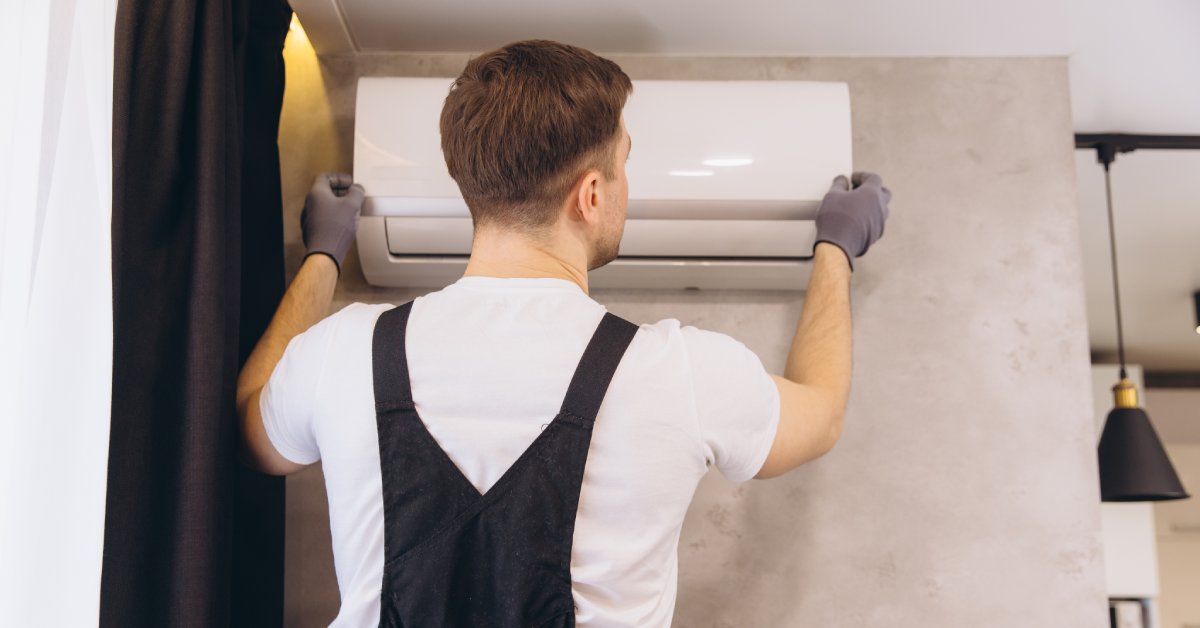 The back side of a man in gray gloves holding both sides of a white mini-split air conditioning unit against a wall.