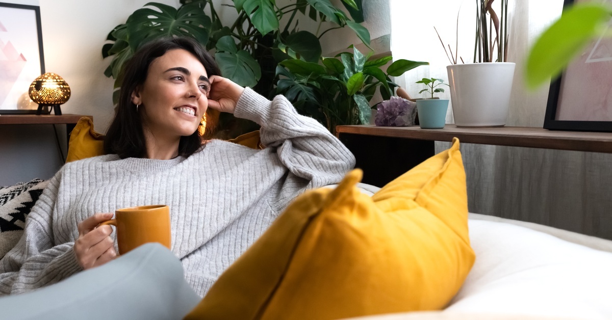 A woman in a light gray sweater surrounded by plants sits on the couch and holds a yellow mug in one hand.