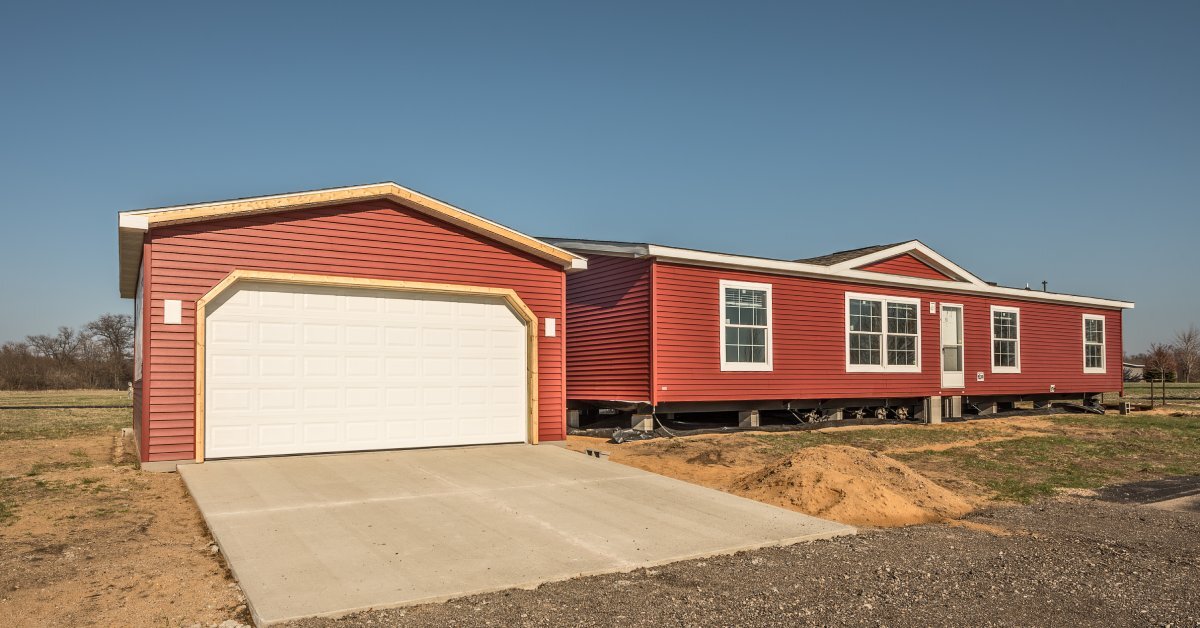 A red manufactured sitting on cinder blocks outside beside a detached, residential garage with matching red siding.
